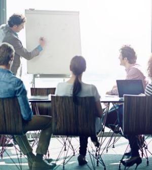 Group of business people sitting at a training room conference table looking at man at easel.